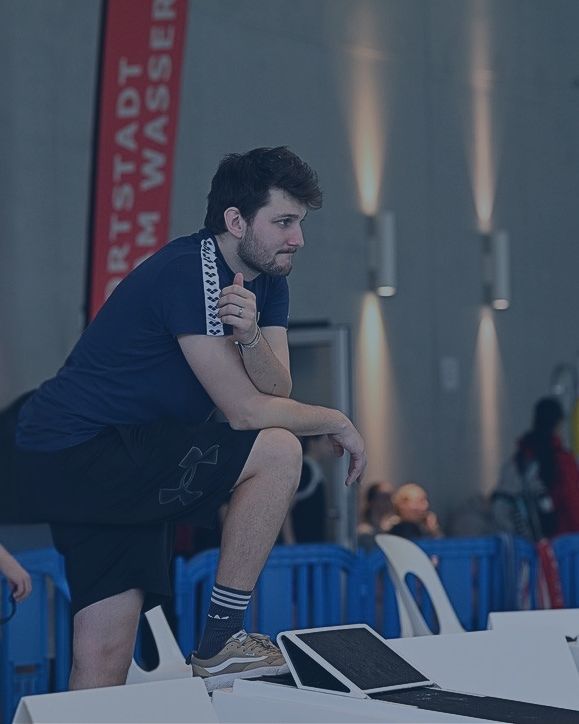 Baptiste Marmoud, accoudé à son genou, suit une compétition de natation au bord du bassin.