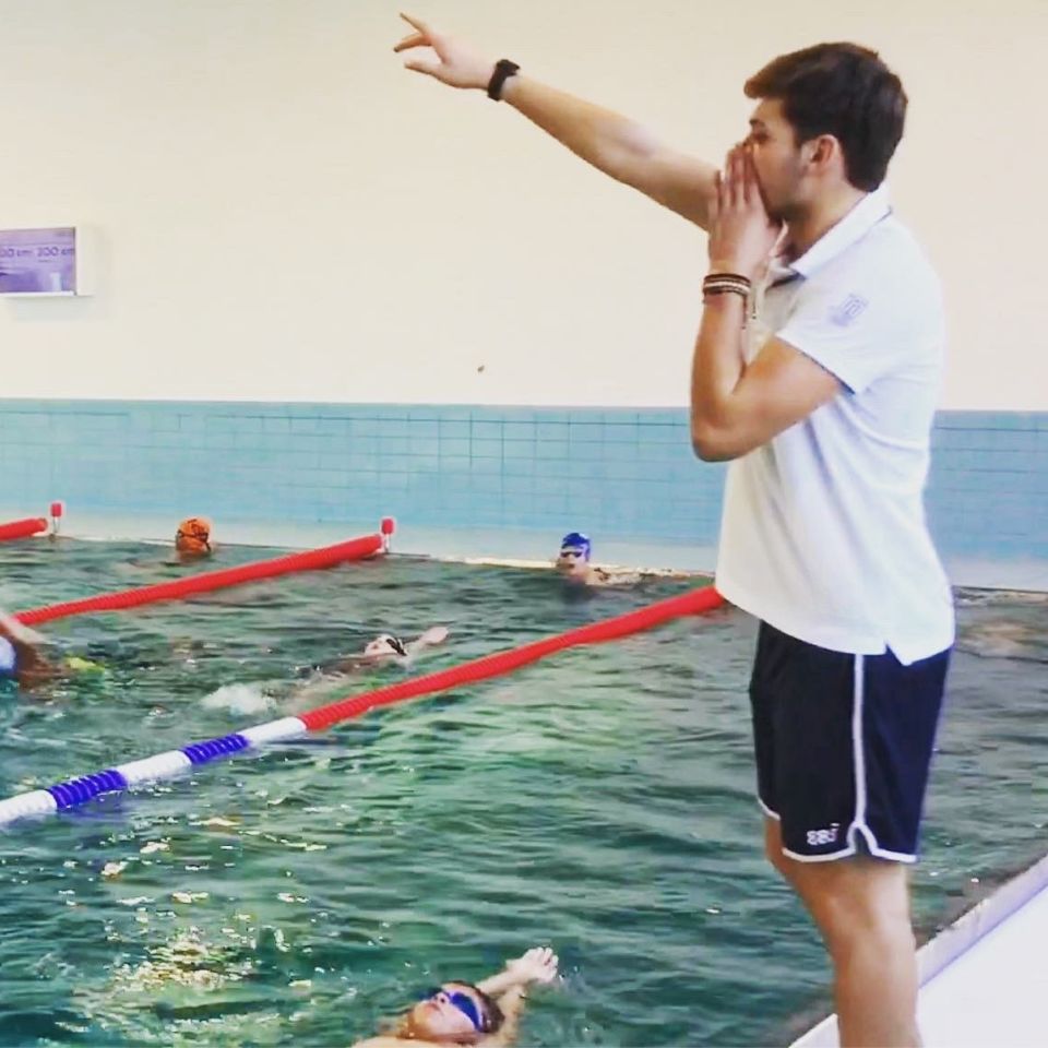 Debout au bord du bassin, Baptiste Marmoud donne une consigne à des nageurs, le bras tendu vers la ligne d'eau.