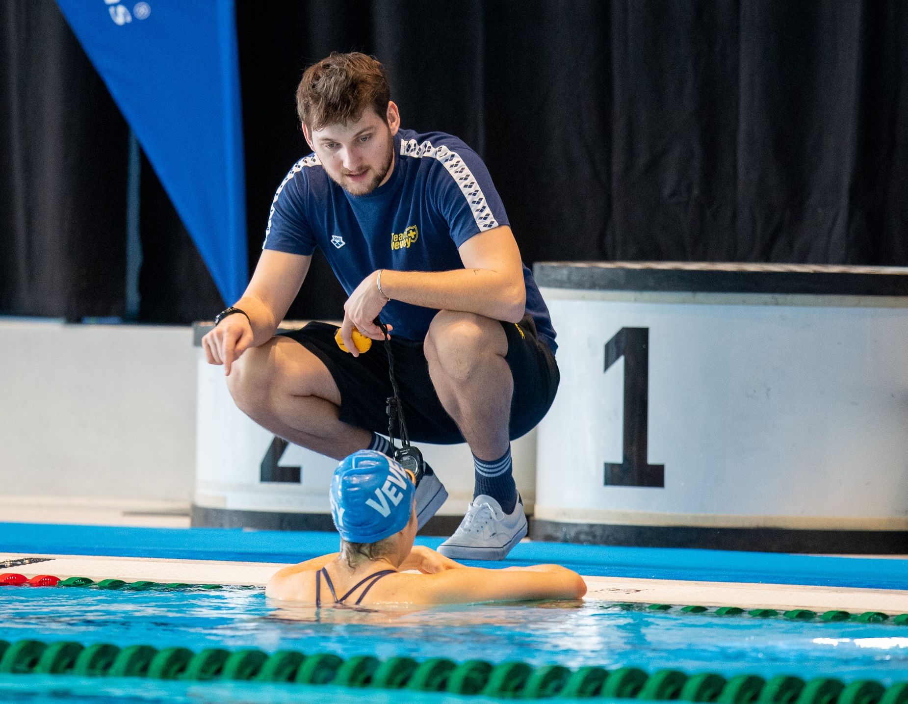 Accroupi près du plot de départ n° 1, Baptiste Marmoud échange avec une nageuse accoudée au bord du bassin.