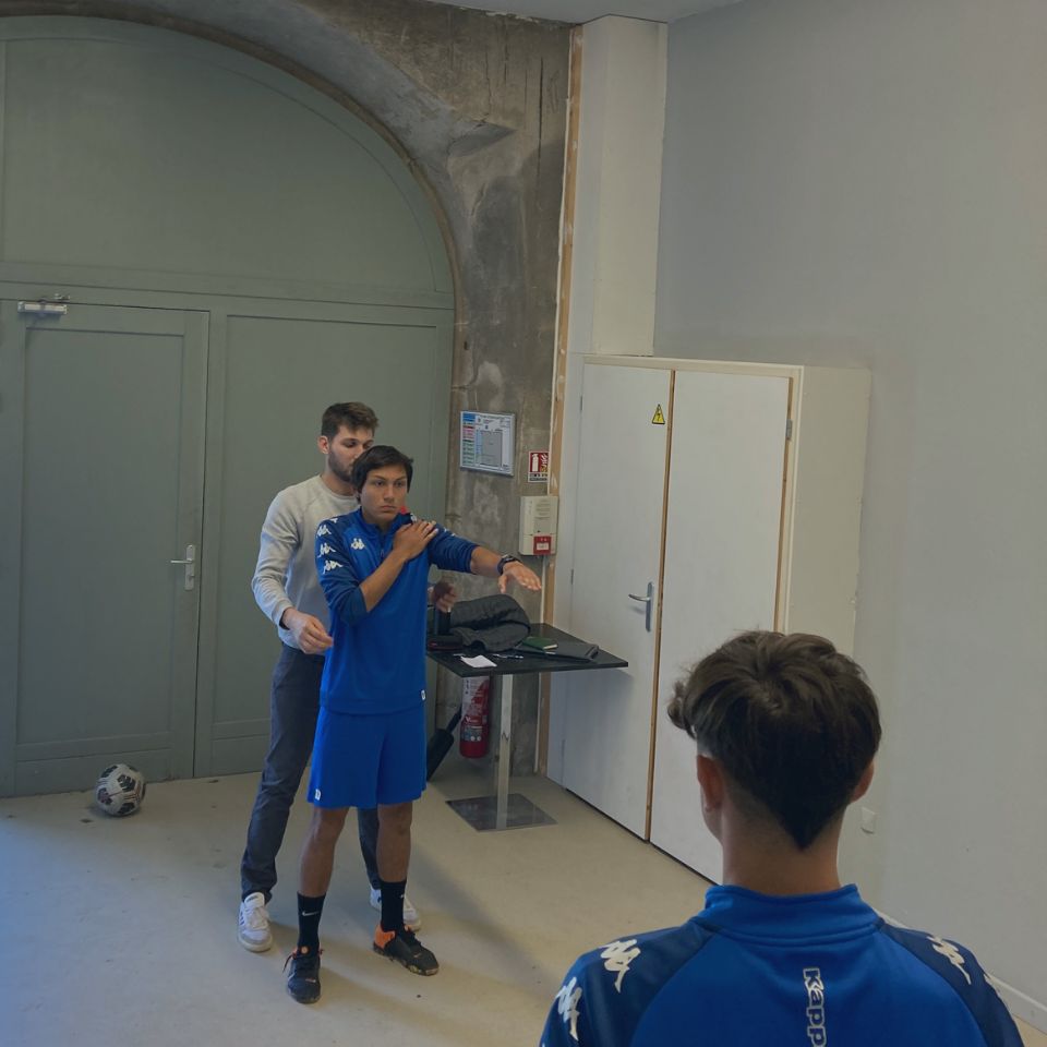 Baptiste Marmoud corrige la posture d'un jeune sportif, bras tendus, sous le regard d'un coéquipier assis.